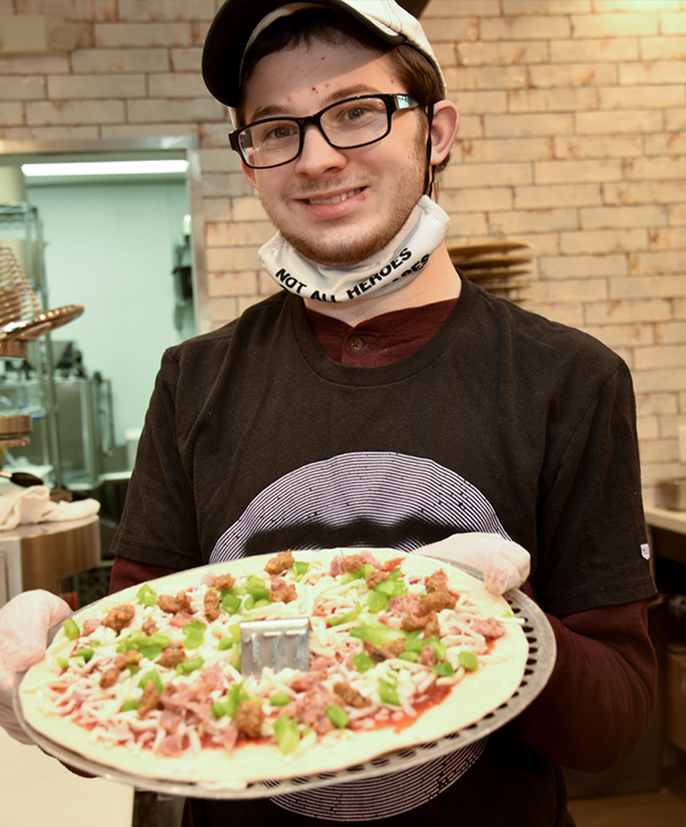 Asaph smiling and holding a pizza in a kitchen.