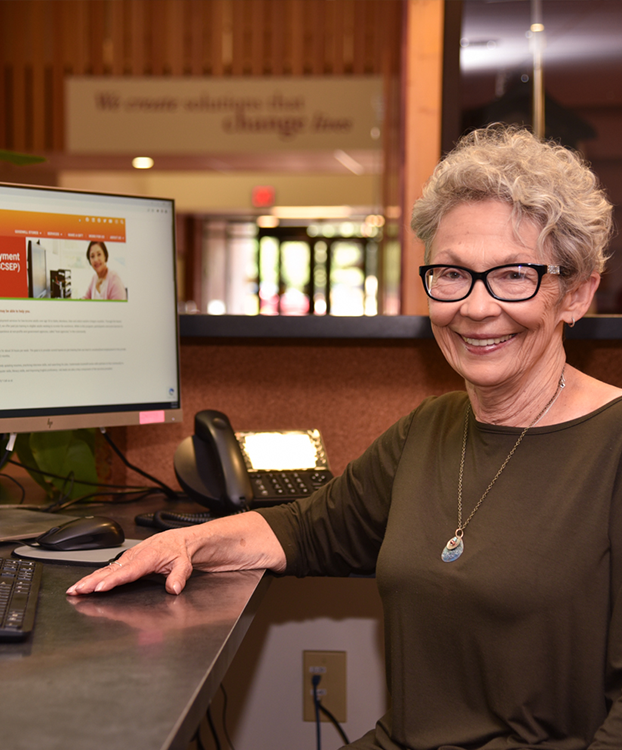 A lady in a green shirt with glasses at a computer.